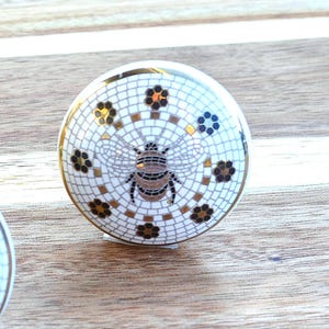 May include: A decorative cabinet knob with a white mosaic pattern, gold accents, and a bee design. The knob has a gold rim and is set against a light wood surface. The bee is brown and black.