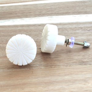 May include: Two white, decorative cabinet knobs with a carved sunburst design. One knob is lying flat, while the other is upright, showing the screw and hardware. The knobs are on a wooden surface.