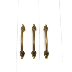 May include: Three matching drawer pulls in an antique brass finish. Each handle features a central fluted bar with decorative leaf-shaped ends at the top and bottom. The hardware has a warm, metallic tone with subtle dark highlights in the crevices.