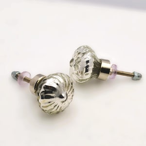 May include: Two silver glass cabinet knobs with a textured, ridged design. The knobs have a metallic finish and are mounted on silver hardware with a pink spacer and a nut. These decorative knobs are suitable for furniture and home decor projects.