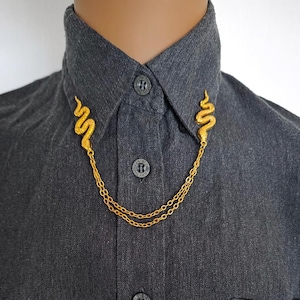 May include: A dark gray button-down shirt featuring a gold-toned collar pin accessory. The accessory consists of two snake-shaped metal pieces attached to each side of the collar, connected by two delicate, draped gold-toned metal chains.