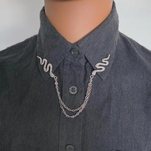 May include: A dark gray button-down shirt featuring a silver-toned collar pin accessory. The accessory consists of two snake-shaped metal pieces attached to each side of the collar, connected by two draped silver chains across the front placket.