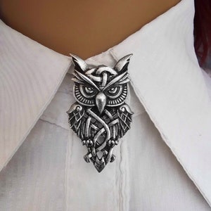 May include: A silver owl brooch featuring an intricate Celtic knotwork design. The owl is facing forward with its wings spread out.