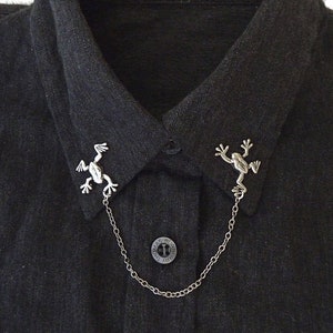 May include: A black collared shirt with a silver chain and two frog-shaped collar clips. The shirt has a label that reads "Thomas Burberry".