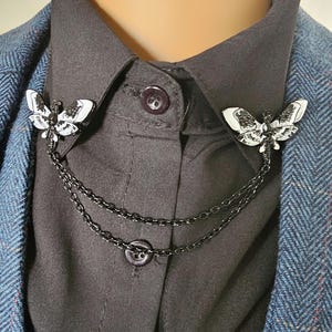 K&ouml;nnte beinhalten: Schwarze und wei&szlig;e Mottenbrosche mit Kette, die die beiden Mottennadeln verbindet. Die Brosche ist an einem schwarzen Hemdkragen und einem blauen Tweed-Blazer befestigt. Die Mottennadeln haben detaillierte Fl&uuml;gelmuster.