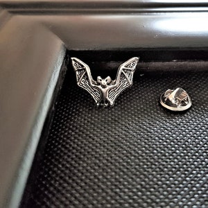 May include: A silver-toned bat pin with a detailed design. The pin is on a black background and includes a separate pin back.