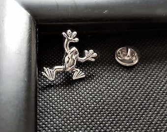May include: A silver frog pin with a textured surface. The pin is attached to a black background.