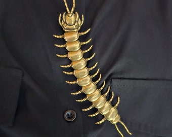 May include: A gold-colored centipede brooch with a detailed design. The brooch is pinned to a black fabric background.