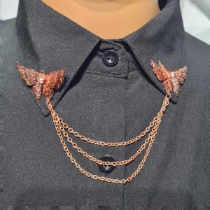 May include: A gold-toned butterfly collar pin with a chain. The butterfly is detailed with a delicate, lacy design. The chain is made of three strands of linked metal.