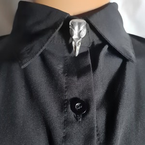 Peut inclure: Une chemise en satin noir avec une épingle en forme de crâne argenté sur le col.