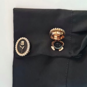 May include: Two gold-toned cufflinks with pearl accents. One cufflink features a black and gold rose design, while the other is a round button with a black centre.