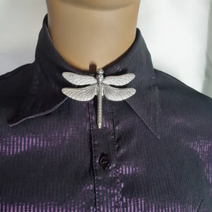 May include: A silver dragonfly brooch pinned to a black and purple striped shirt collar.