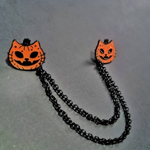 May include: A black chain necklace with two orange pumpkin-shaped cat face charms. The charms have black eyes and a black mouth.