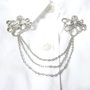 May include: A silver octopus-shaped collar pin with three chains attached. The pin is on a white shirt.