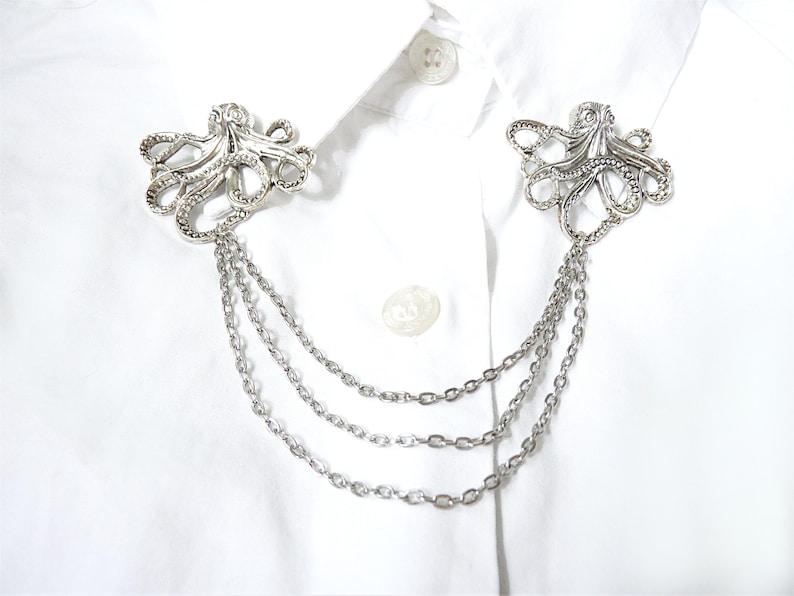 May include: A silver octopus-shaped collar pin with three chains attached. The pin is on a white shirt.