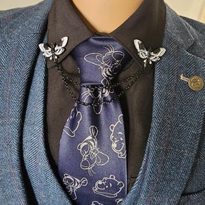 K&ouml;nnte beinhalten: Eine marineblaue Krawatte mit wei&szlig;em Winnie-Puuh-Muster wird von zwei schmetterlingsf&ouml;rmigen Broschen gehalten. Die Broschen sind durch eine Kette verbunden und an einem schwarzen Hemd befestigt. Das Ensemble wird durch einen blauen Fischgr&auml;tenanzug erg&auml;nzt.