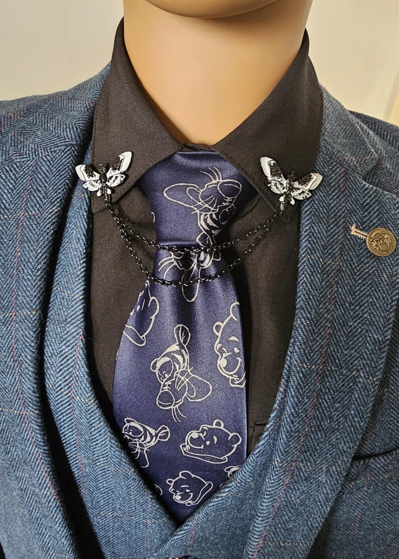 K&ouml;nnte beinhalten: Eine marineblaue Krawatte mit wei&szlig;em Winnie-Puuh-Muster wird von zwei schmetterlingsf&ouml;rmigen Broschen gehalten. Die Broschen sind durch eine Kette verbunden und an einem schwarzen Hemd befestigt. Das Ensemble wird durch einen blauen Fischgr&auml;tenanzug erg&auml;nzt.