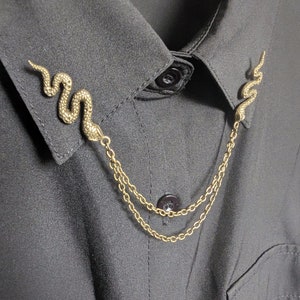 Könnte beinhalten: Goldfarbene Schlangen-Kragenclips mit einer Doppel-Kette, befestigt an einem schwarzen Hemd mit Kragen. Die Schlangenclips sind mit Schuppen und einem geschwungenen Design versehen. Das Hemd hat einen Knopfverschluss.