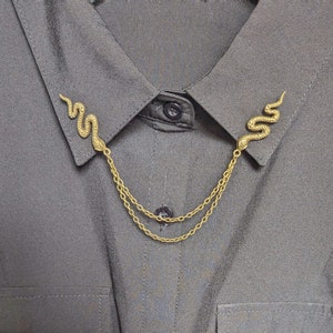 May include: Gold-colored snake collar clips connected by delicate chains. The snake-shaped clips are attached to the collar of a gray shirt. The jewelry adds a unique and stylish accent to the shirt.