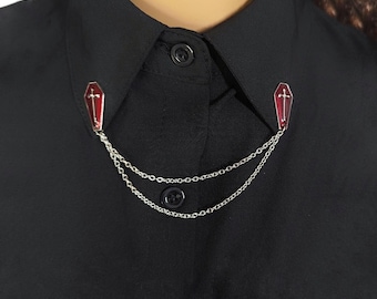 Può includere: Una coppia di spille da colletto a forma di bara in rosso scuro con croci argentate, collegate da due delicate catenelle metalliche color argento. Le spille sono fissate alle punte di una camicia nera, creando un collegamento decorativo.