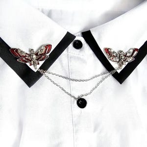 May include: A white collared shirt with a black trim and two silver butterfly brooches with red and black accents connected by a silver chain.