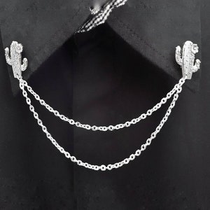May include: A silver chain with two cactus-shaped charms attached to it. The chain is draped over a black fabric background.