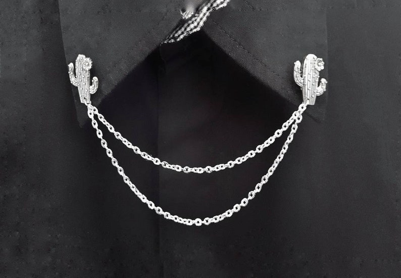May include: A silver chain with two cactus-shaped charms attached to it. The chain is draped over a black fabric background.
