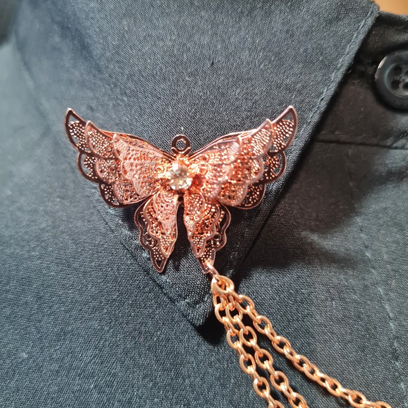May include: A rose gold butterfly brooch with a clear rhinestone in the centre. The brooch has a delicate filigree design and is attached to a chain.