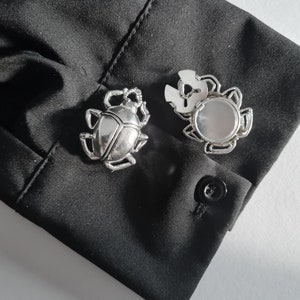 May include: Two silver cufflinks, one shaped like a beetle and the other a circular design with a clear stone in the center.