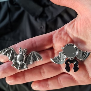 Puede incluir: Dos broches de plata en forma de murciélago. Un broche es un diseño de murciélago sólido, mientras que el otro es un murciélago con bisagras con un espejo en el interior.