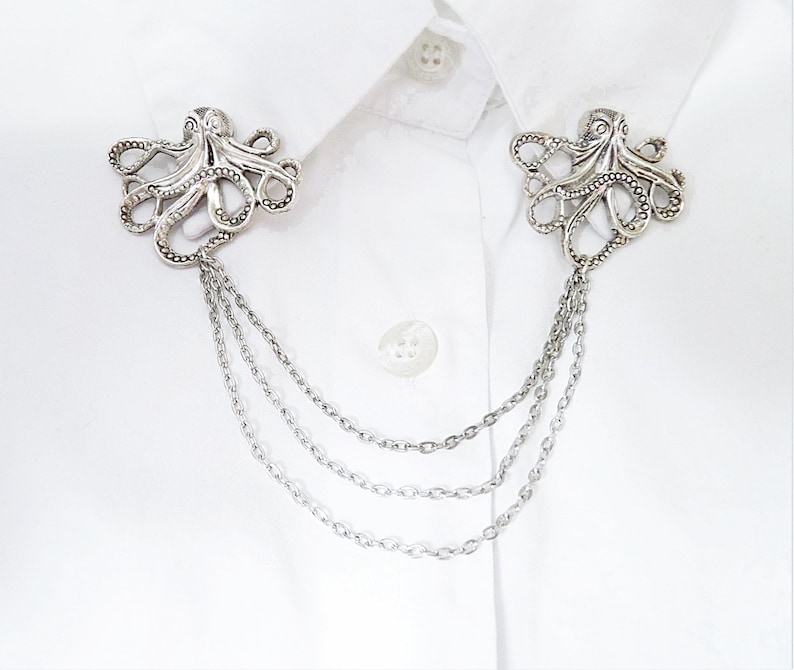 May include: A pair of silver octopus collar clips with a chain attached to each. The clips are on a white shirt.