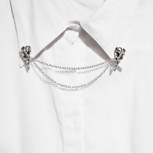 Puede incluir: Un broche de collar plateado con un dise&ntilde;o de calavera y una cadena doble. El broche est&aacute; sujeto a una camisa blanca.