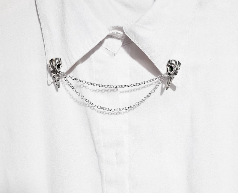Puede incluir: Un broche de collar plateado con un dise&ntilde;o de calavera y una cadena doble. El broche est&aacute; sujeto a una camisa blanca.