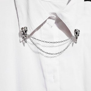 Puede incluir: Un broche de collar de cadena plateada con dos dijes en forma de calavera. El broche est&aacute; sujeto a una camisa blanca.