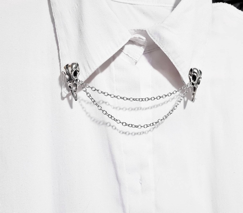 Puede incluir: Un broche de collar de cadena plateada con dos dijes en forma de calavera. El broche est&aacute; sujeto a una camisa blanca.