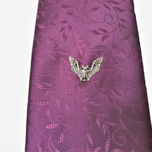 May include: A silver bat tie tack on a purple patterned tie. The tie tack is a detailed, realistic depiction of a bat with its wings spread.