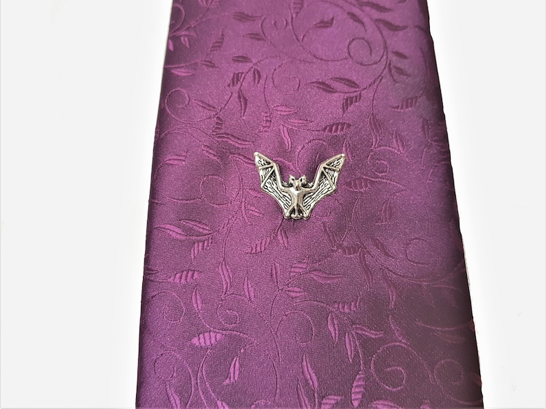 May include: A silver bat tie tack on a purple patterned tie. The tie tack is a detailed, realistic depiction of a bat with its wings spread.