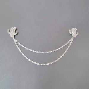 May include: A silver chain necklace with two cactus charms. The charms are made of silver and have a sparkly finish.