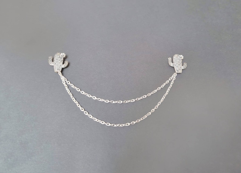 May include: A silver chain necklace with two cactus charms. The charms are made of silver and have a sparkly finish.