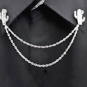May include: A silver chain with two cactus-shaped charms attached to the ends. The chain is draped over a black shirt collar.