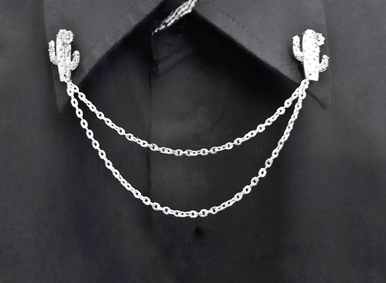 May include: A silver chain with two cactus-shaped charms attached to the ends. The chain is draped over a black shirt collar.