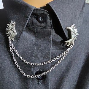 Può includere: Una collana a catena d'argento con due ciondoli, uno con un sole e l'altro con una luna crescente. I ciondoli sono collegati alla catena con un piccolo anello.