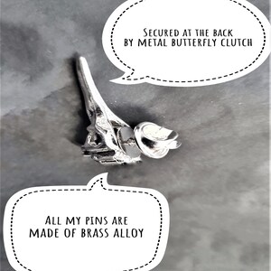 May include: A silver metal pin shaped like the Eiffel Tower with a butterfly clasp on the back. The pin is made of brass alloy. "SECURED AT THE BACK BY METAL BUTTERFLY CLUTCH" and "ALL MY PINS ARE MADE OF BRASS ALLOY" are written in black text on white speech bubbles.