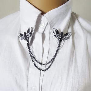 K&ouml;nnte beinhalten: Schwarze und wei&szlig;e Motten-Kragennadel mit Doppelketten, getragen auf einem wei&szlig;en Button-Down-Hemd. Die Mottennadeln sind mit detaillierten Fl&uuml;gelmustern versehen. Die Kette hat ein dunkles, mattes Finish.