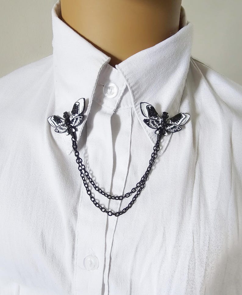 K&ouml;nnte beinhalten: Schwarze und wei&szlig;e Motten-Kragennadel mit Doppelketten, getragen auf einem wei&szlig;en Button-Down-Hemd. Die Mottennadeln sind mit detaillierten Fl&uuml;gelmustern versehen. Die Kette hat ein dunkles, mattes Finish.