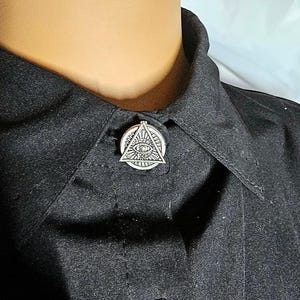 May include: A silver-toned metal pin featuring an eye of providence design, which consists of an eye centered within a triangle, attached to the collar of a black button-down shirt. The pin has a circular base with a textured, gear-like edge.
