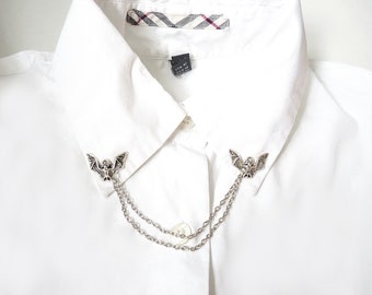 Può includere: Una camicia bianca con colletto e un fermaglio per colletto a catena argentata con due disegni di pipistrelli.