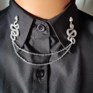 May include: A silver snake collar pin with a double chain. The pin is attached to a black shirt.