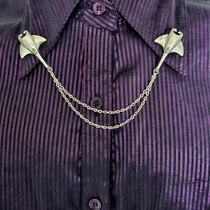 May include: A silver-toned collar pin set featuring two stingray-shaped charms connected by a double-strand chain. The metallic stingrays are attached to the points of a dark purple and black striped button-down shirt collar with a central button.
