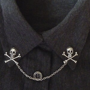 May include: A pair of silver skull and crossbones collar clips connected by a silver chain. The clips are attached to a black collared shirt.